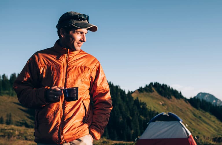 Traveler in an orange jacket holding a mug near a tent in the mountains